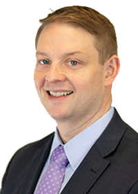 A man with short blond hair wearing a charcoal suit and purple tie smiles at the camera. 