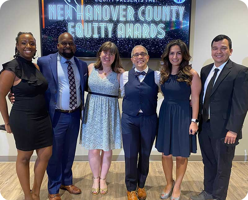 A diverse group of people pose together in front of the New Hanover County Equity Awards sign. 