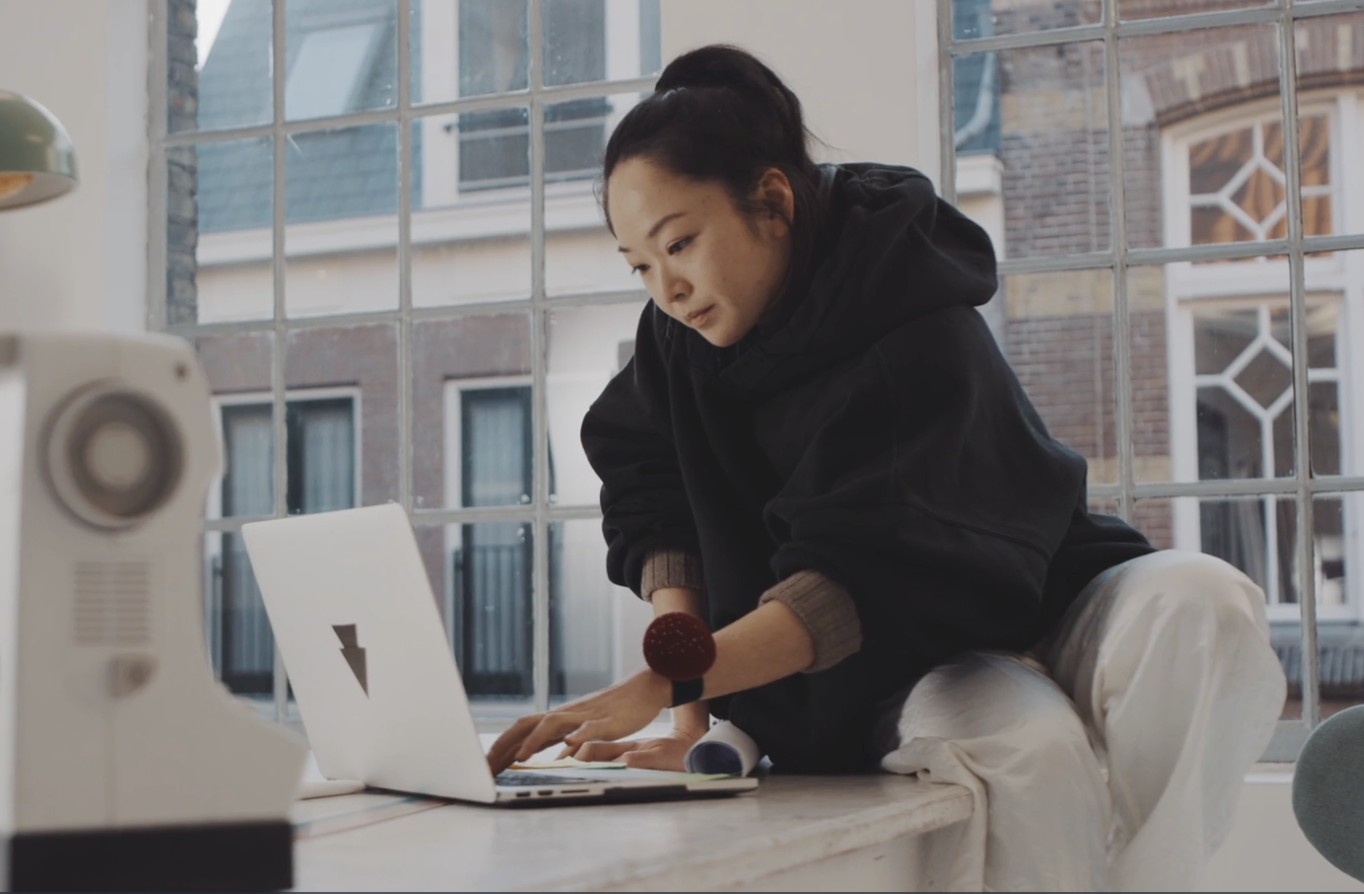 A wants in a dark hoodie sits at a table as she works on her laptop in her studio. 